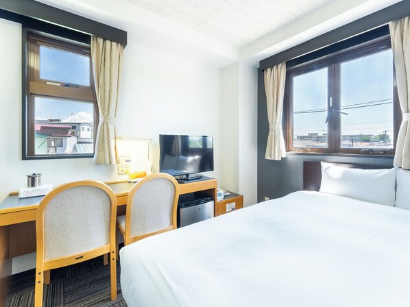 Et hotelværelse med en seng, et skrivebord med tv, to stole og vinduer med gardiner.