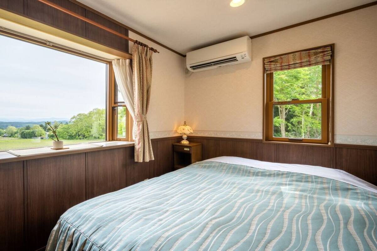 A bedroom with a large window, a bed with a patterned bedspread, a bedside table with a lamp, and an air conditioning unit on the wall.