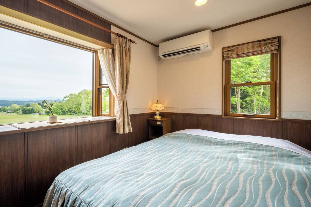A bedroom with a bed, a nightstand, an air conditioner, a window with curtains, and a view of greenery.