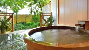 Zona de baños termales con una bañera de madera, rodeada de vegetación y una valla de madera.