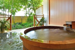 Zona de baños termales con una bañera de madera, rodeada de vegetación y una valla de madera.