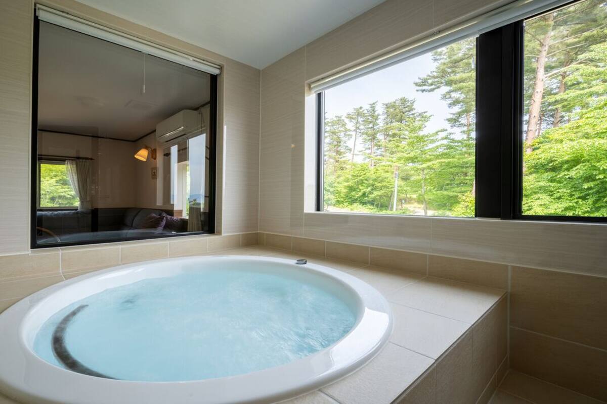A modern bathroom with a large bathtub, a window with a forest view, and a mirror reflecting the room.