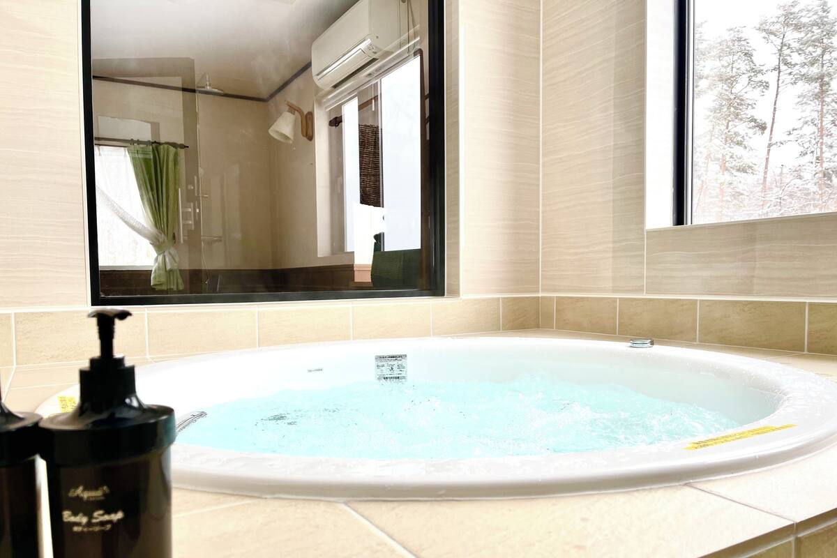 A bathroom with a bathtub, a window with a view of trees, and a bottle of body soap on the counter.
