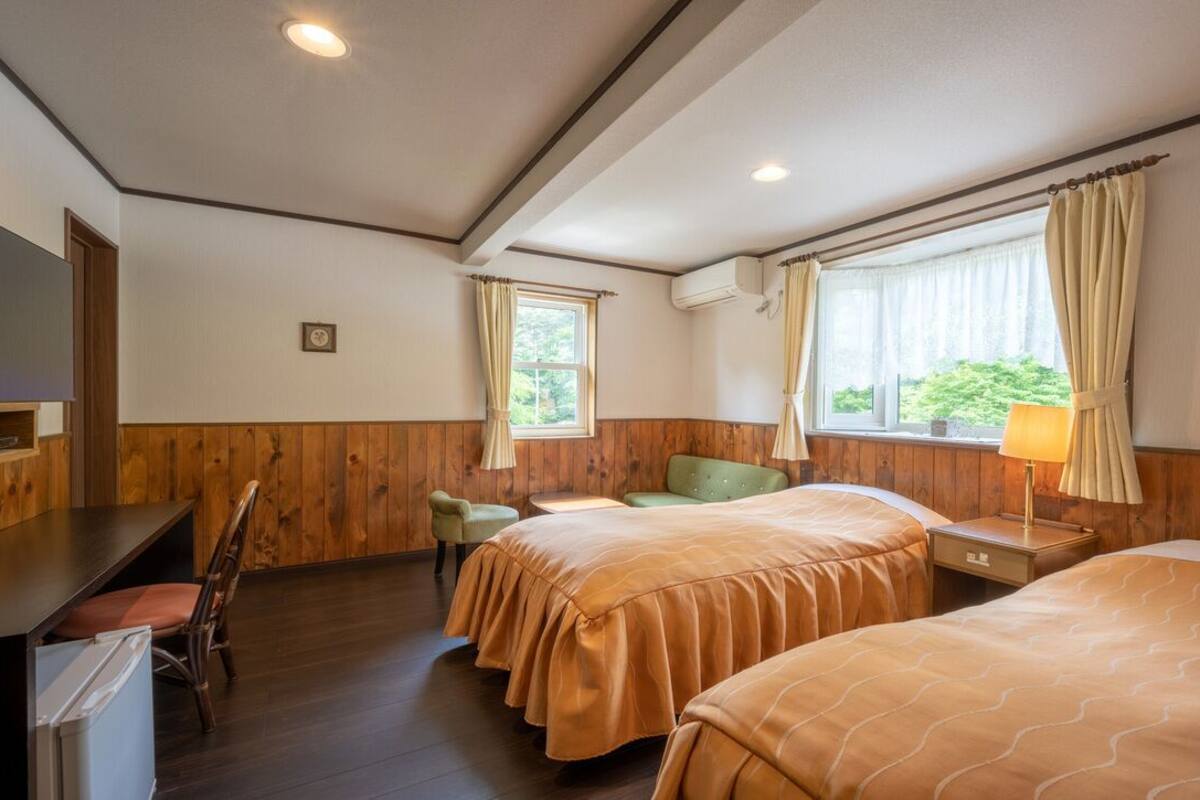 A hotel room with two beds, a desk, a chair, a window with curtains, and an air conditioning unit.