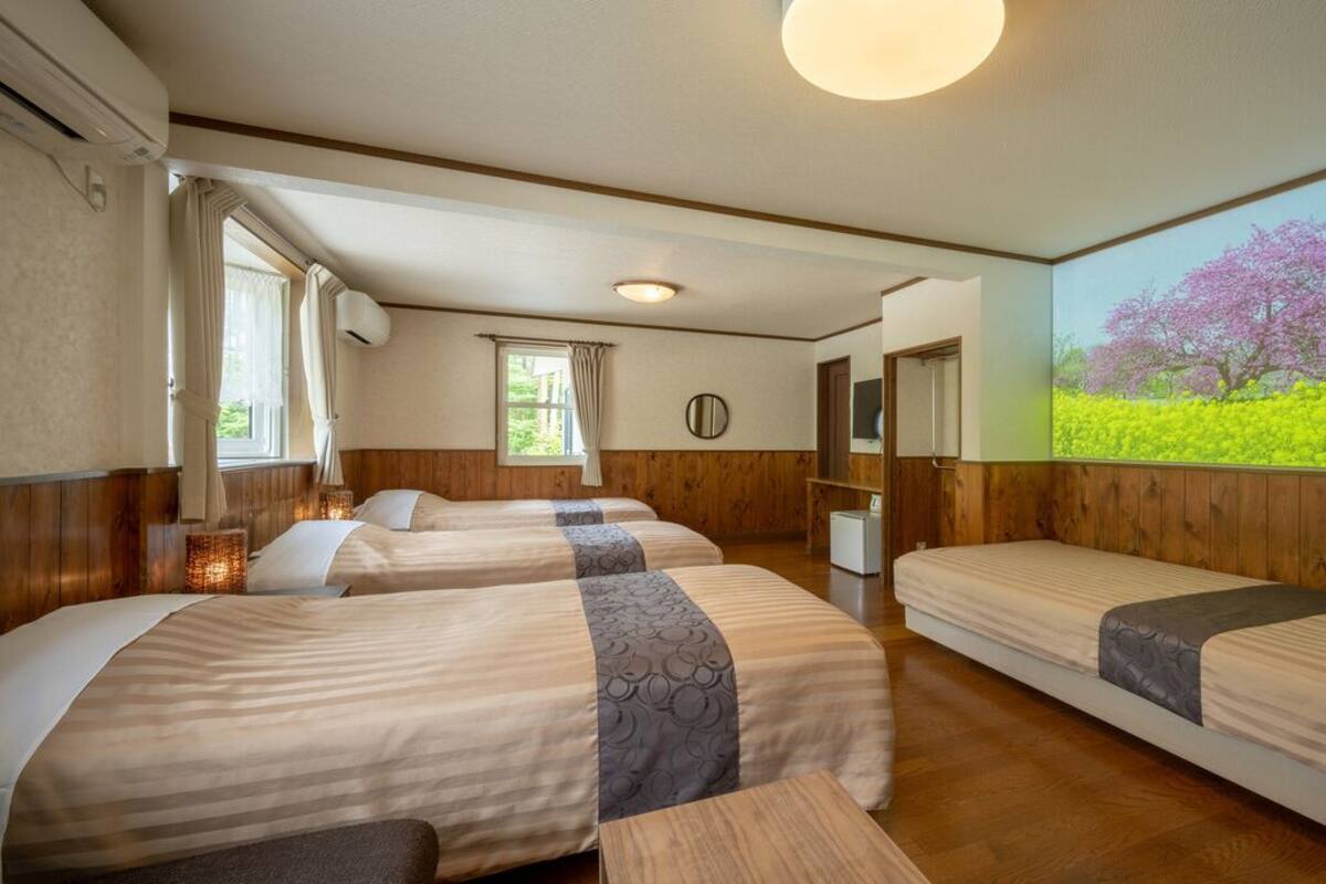 A hotel room with two beds, a wooden floor, a large window with curtains, a wall-mounted TV, and a painting of a tree.