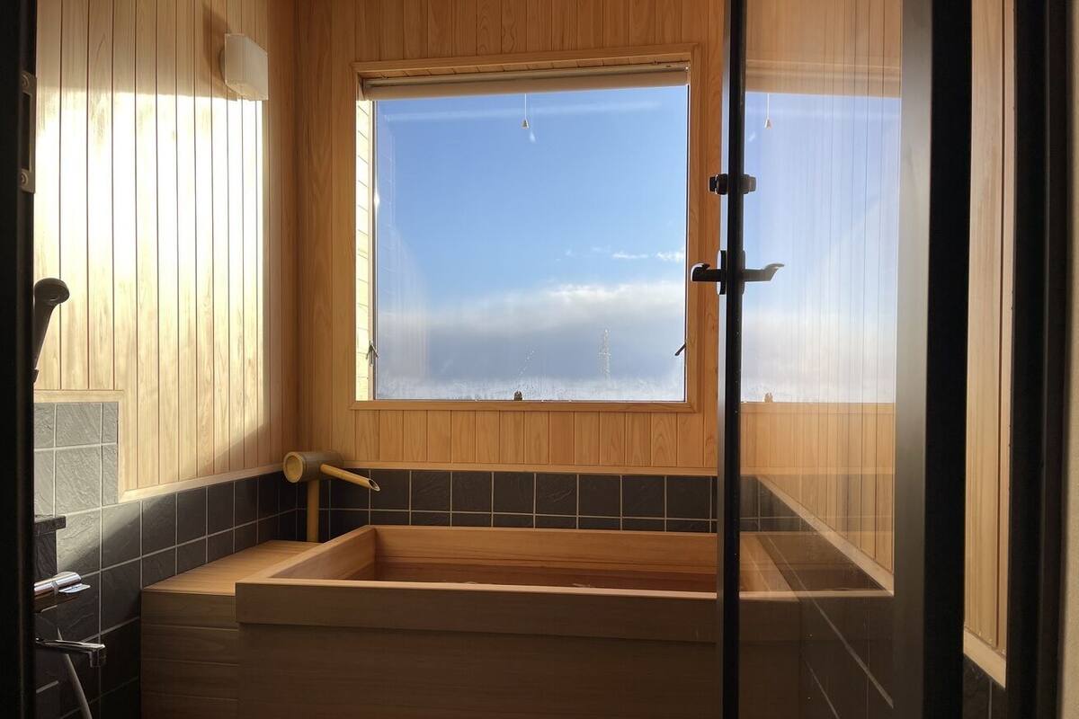 A wooden bathtub with a window, a towel rack, and a shower area.