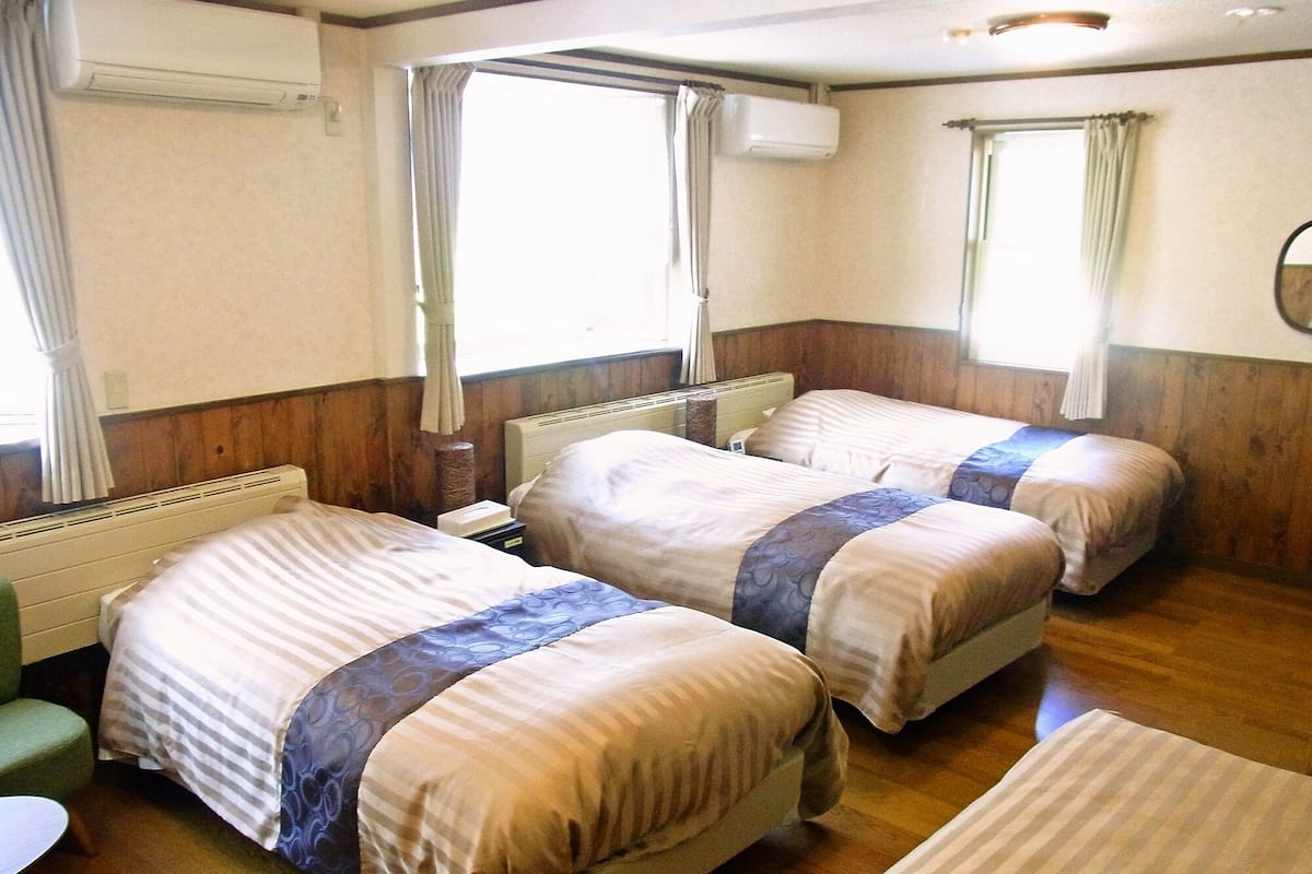 Quarto de hotel com duas camas, um radiador, uma mesa pequena e uma janela com cortinas.