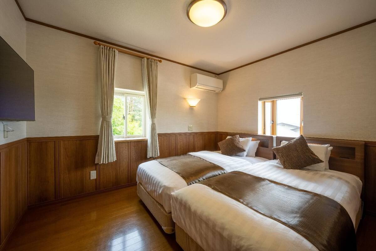 A hotel room with a large bed, a flat-screen TV, an air conditioning unit, and a window with curtains.