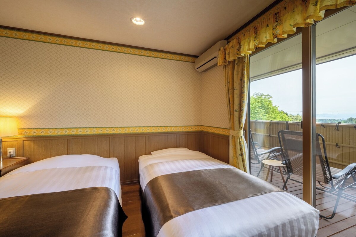 A hotel room with two beds, a window with curtains, a chair, and a view of the outdoors.