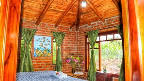 A room with a wooden ceiling, brick walls, a bed with patterned bedding, a painting on the wall, and a window with curtains. - Kalera Lodge - Equator Face (Cotacachi)