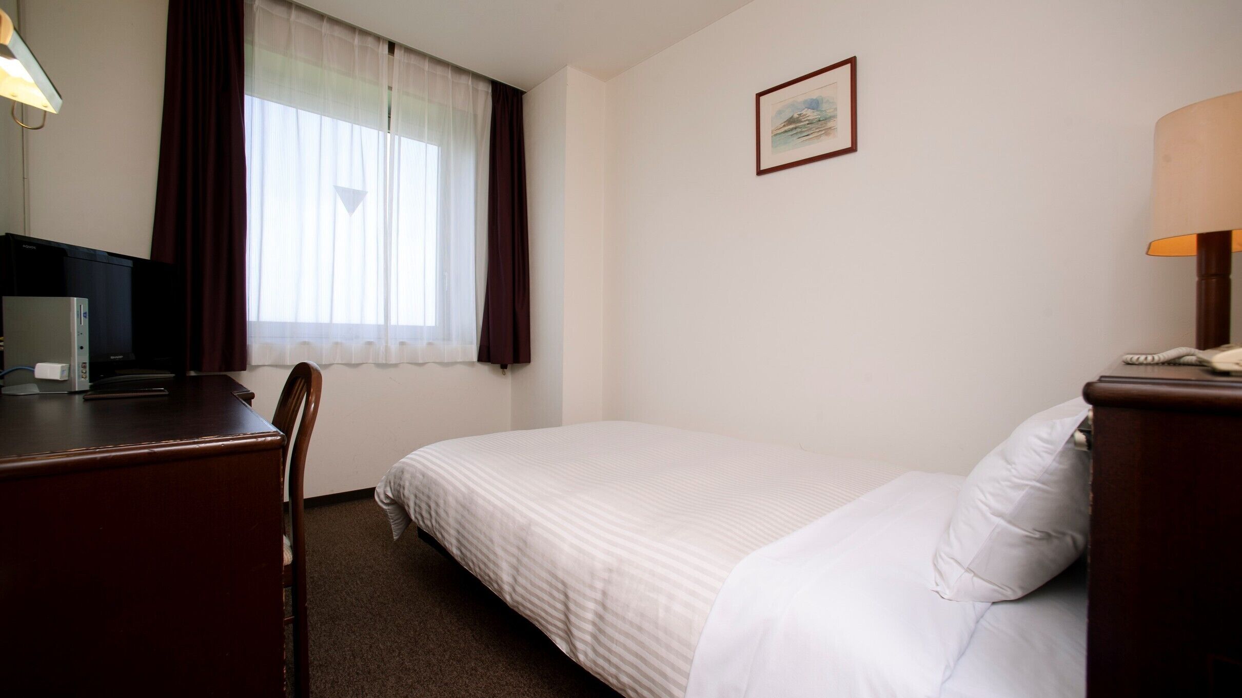 Una habitació d'hotel amb un llit, una taula de nit, una cadira, una làmpada, un quadre a la paret i una finestra amb cortines.