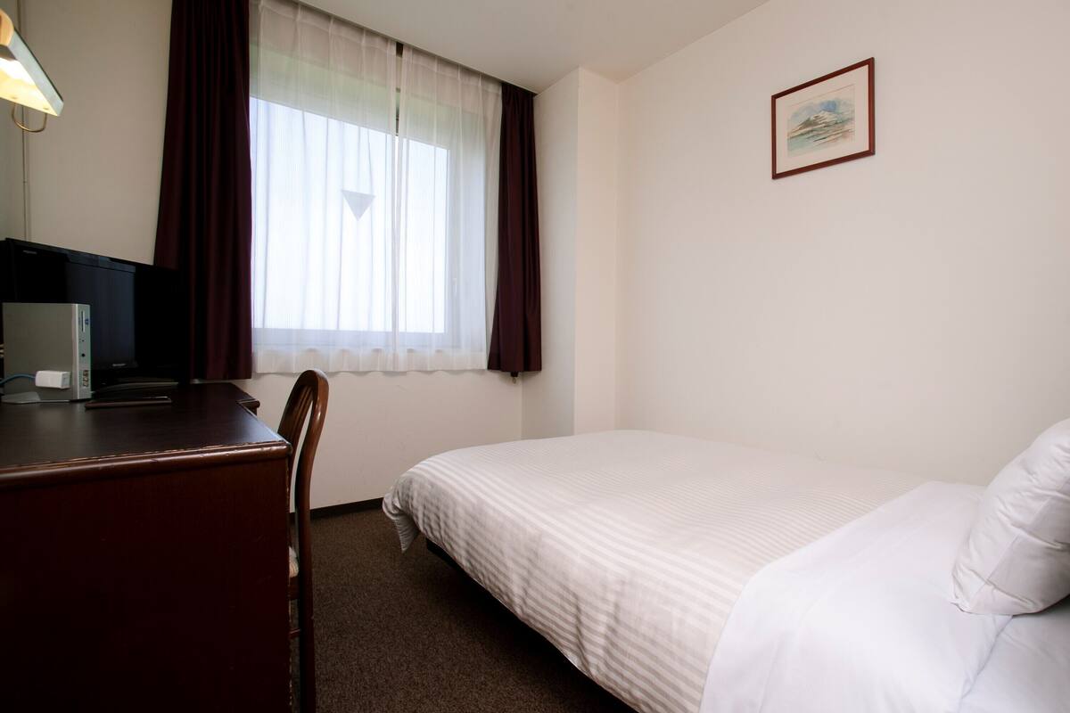 A hotel room with a bed, a desk with a computer, a chair, a window with curtains, a framed picture on the wall, and a bedside lamp.