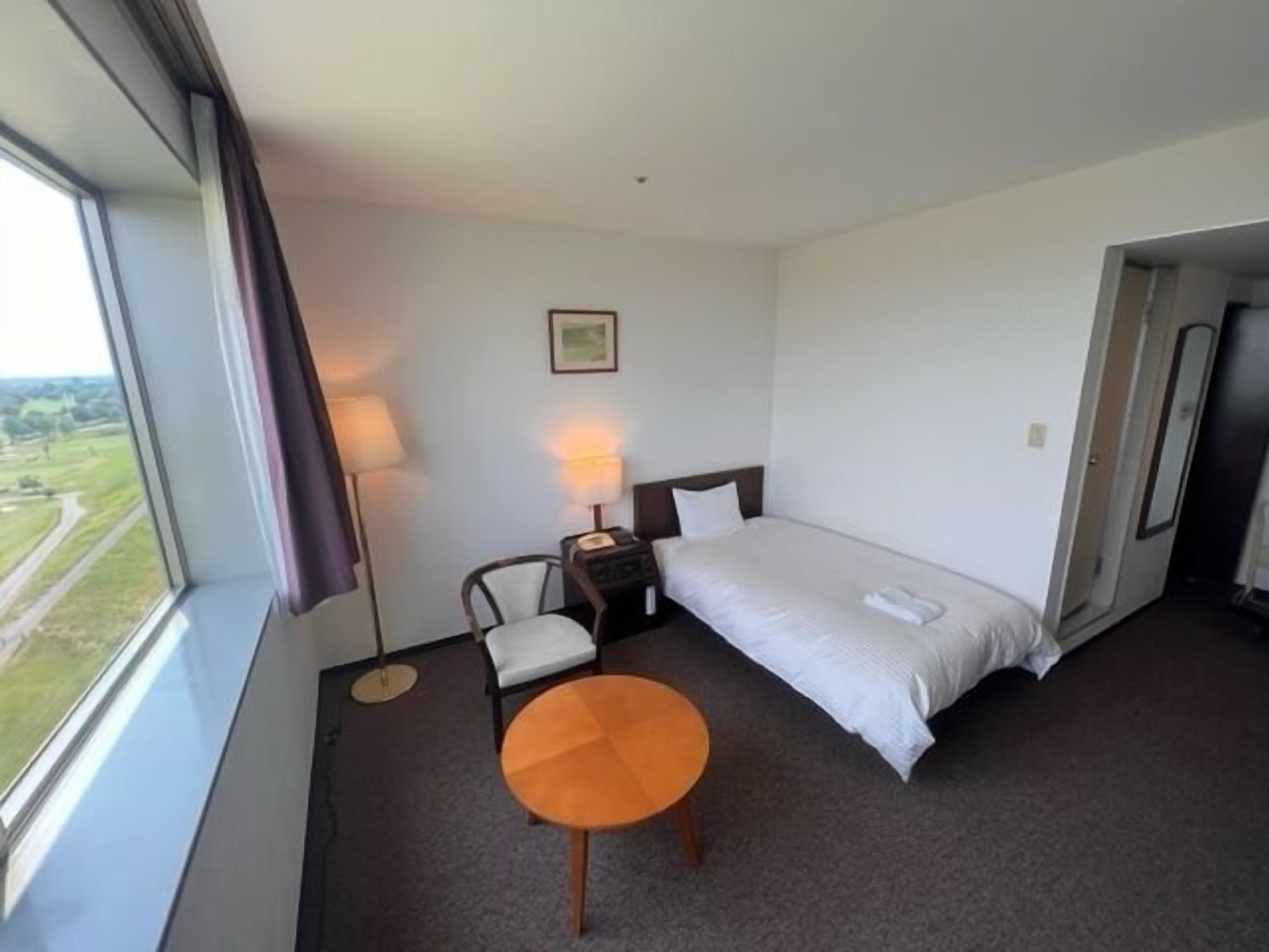 A hotel room with a bed, a chair, a small table, a nightstand, a lamp, and a window with a view.