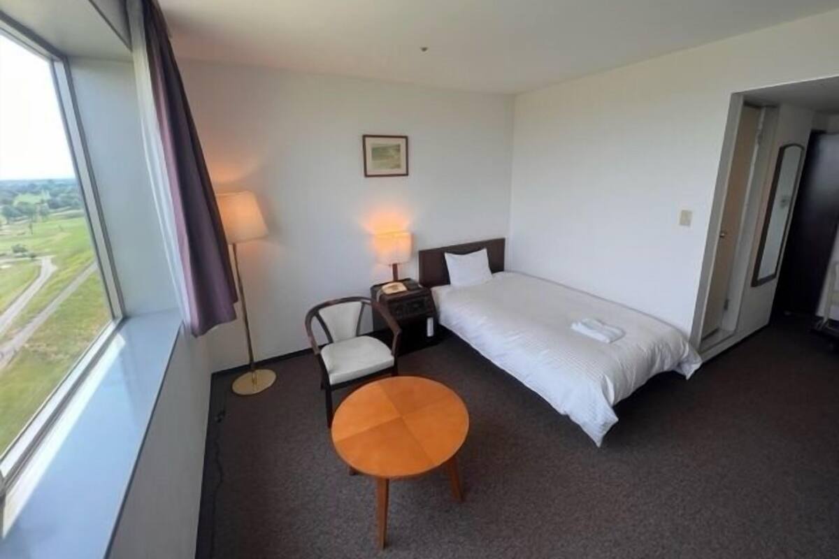 A hotel room with a bed, a chair, a small table, a nightstand, a lamp, and a window with a view.