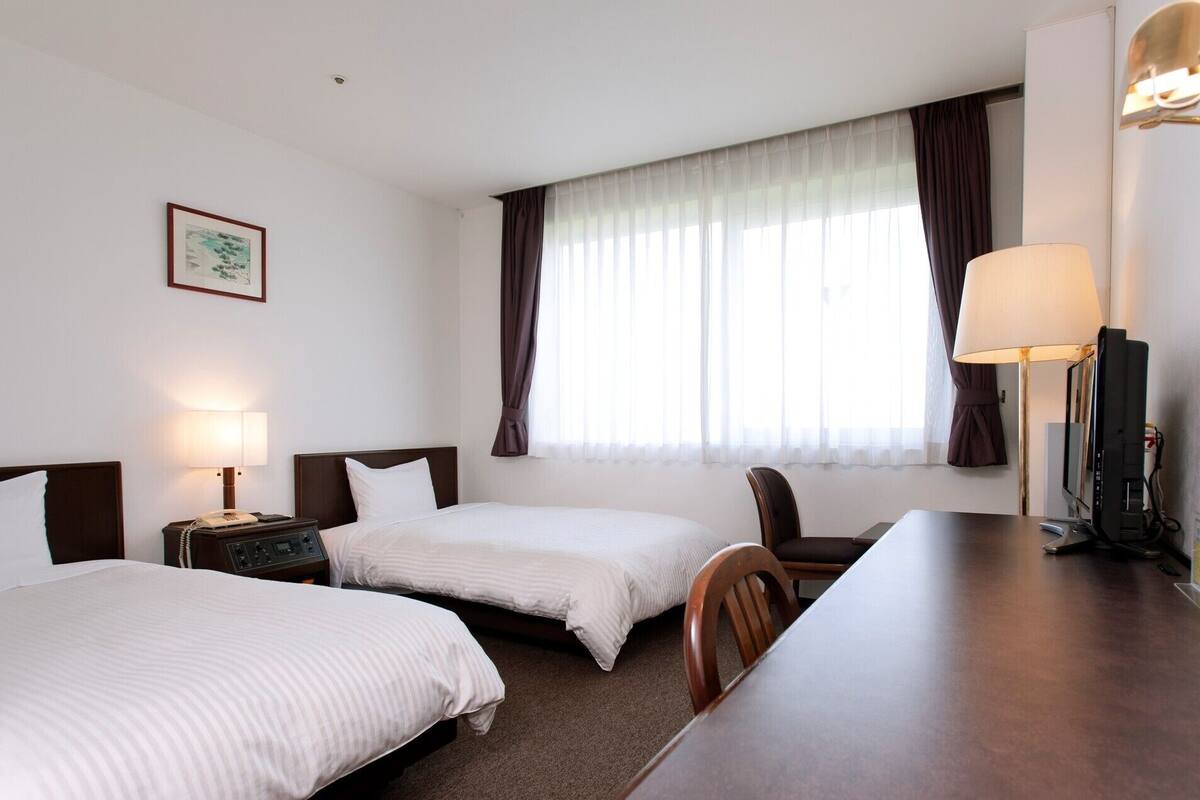 A hotel room with two beds, a desk, a chair, a TV, and a window with curtains.