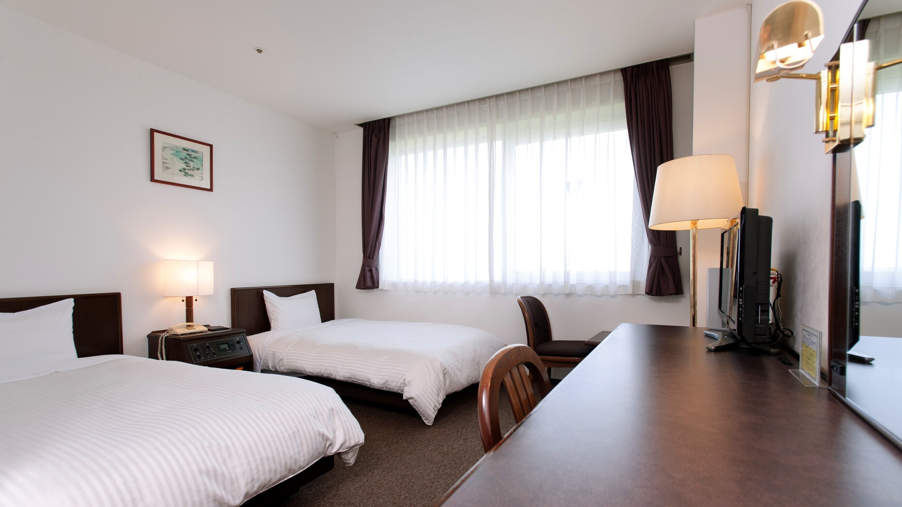 A hotel room with two beds, a desk, a chair, a desk lamp, a television, and a window with curtains.