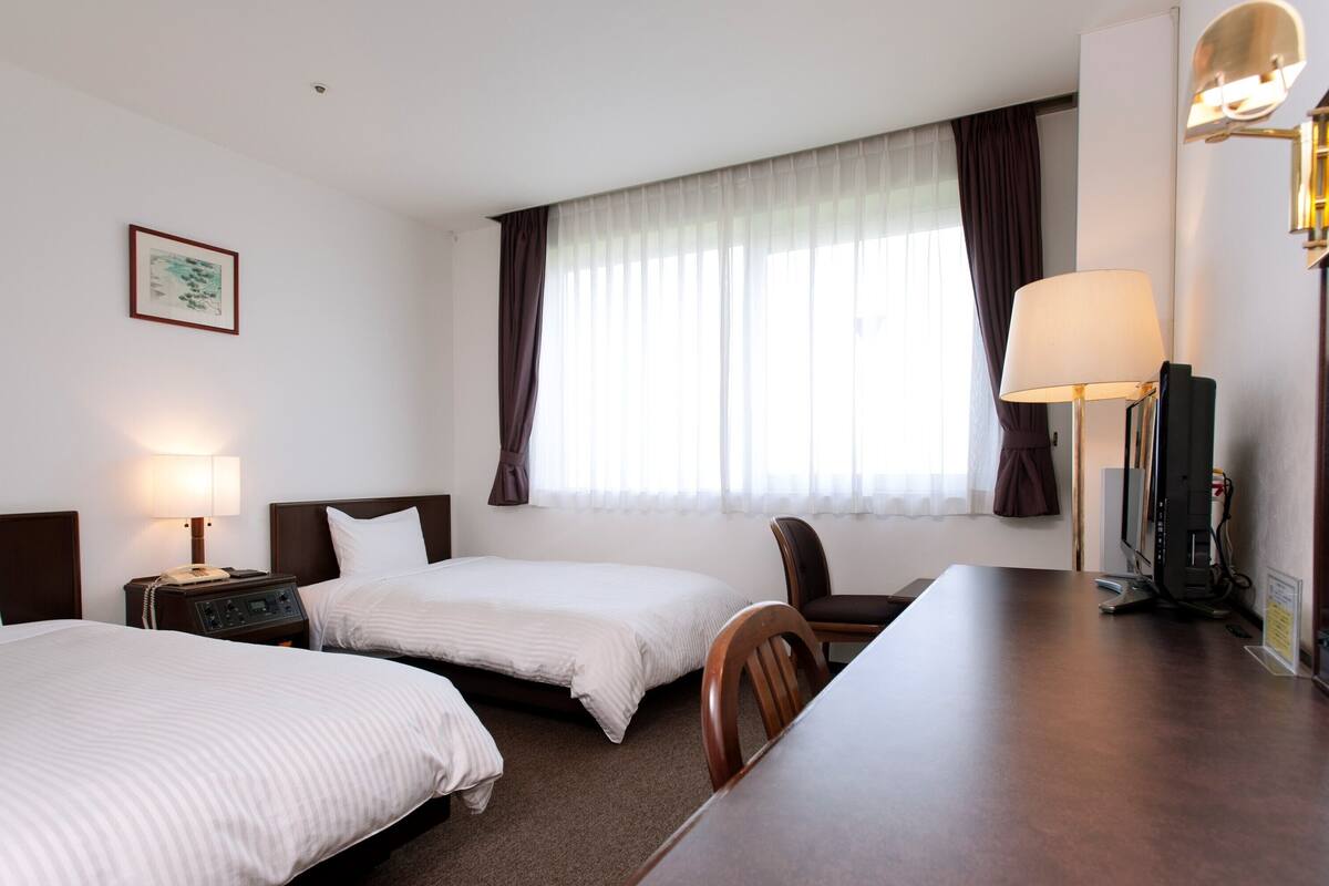 A hotel room with two beds, a desk, a chair, a desk lamp, a television, and a window with curtains.