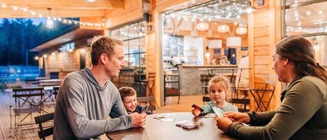 Eine vierköpfige Familie sitzt draußen an einem runden Tisch, mit Holzdeck und Lichterkette darüber.