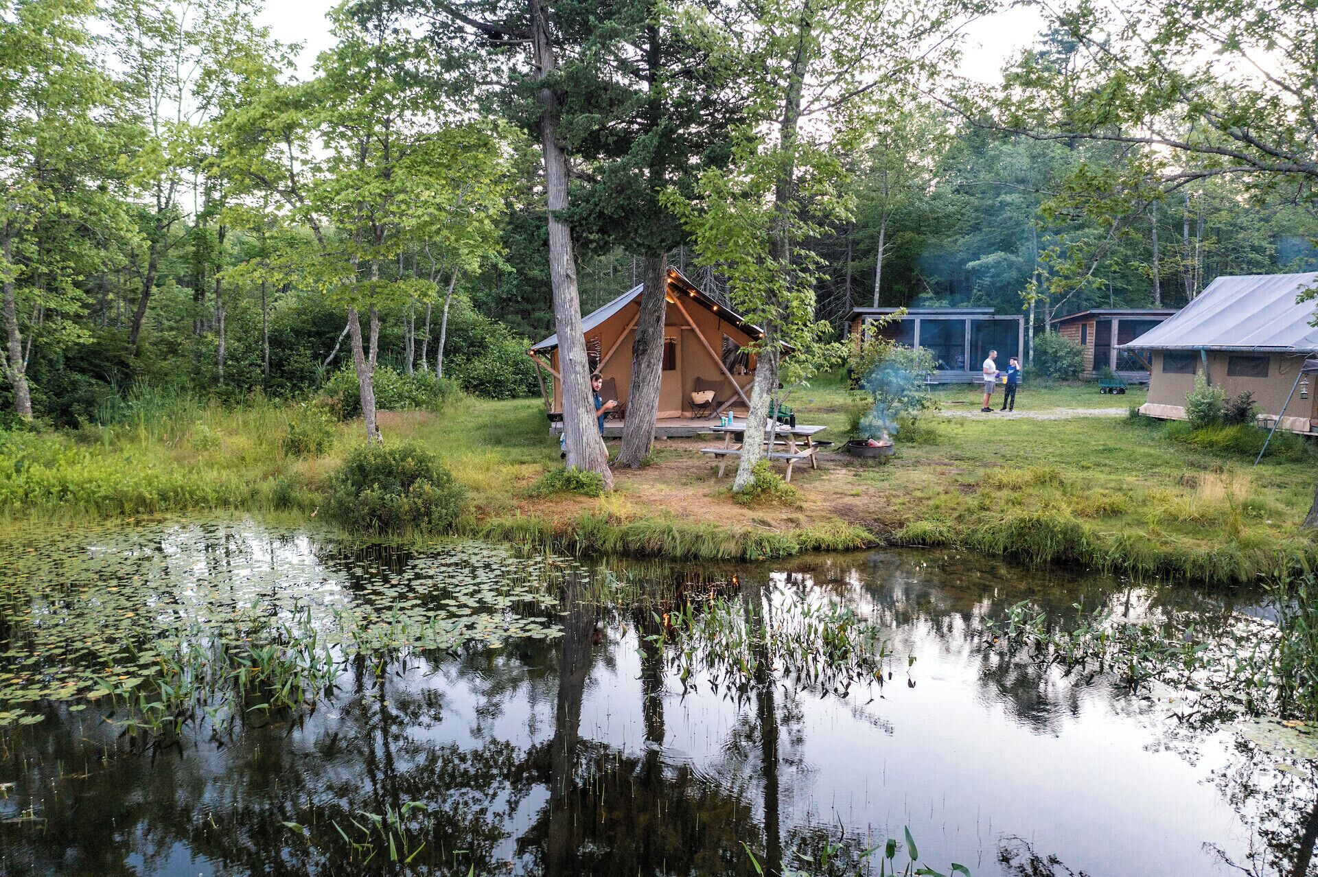A rustic cabin with a deck, surrounded by trees and a pond.