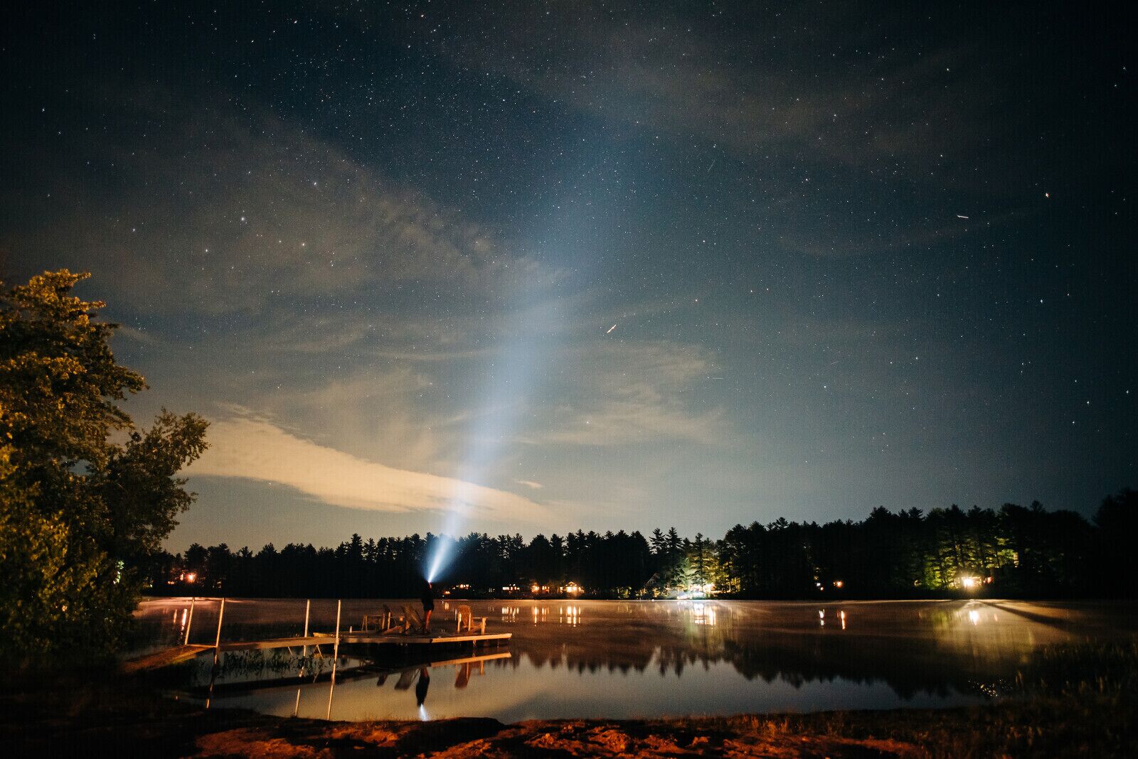 A night sky with stars over a calm body of water, a dock, and a forest in the background.