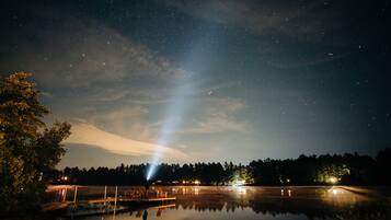 A night sky with stars over a calm body of water, a dock, and a forest in the background.