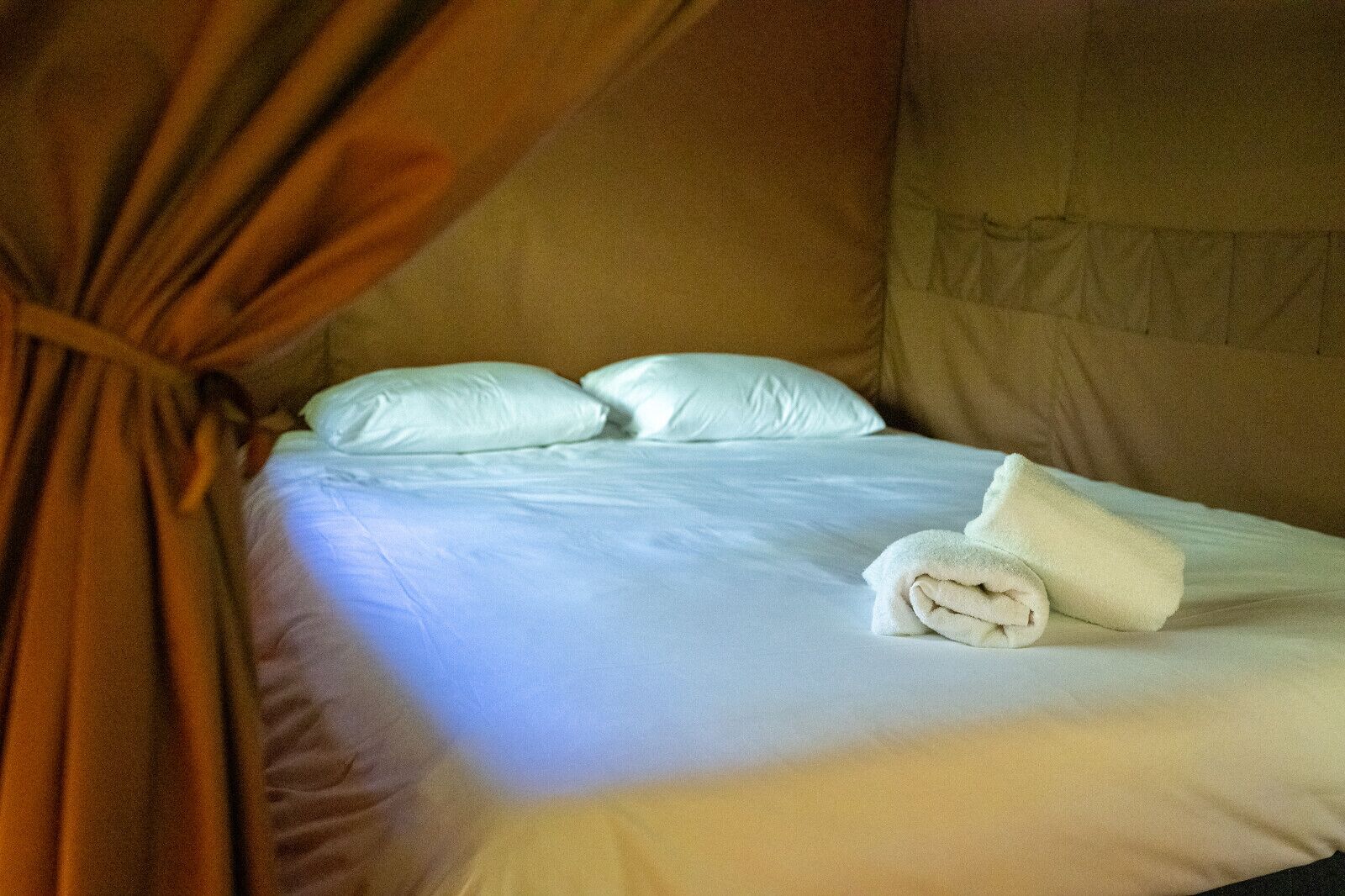 Una cama con sábanas blancas, dos almohadas y una toalla enrollada.