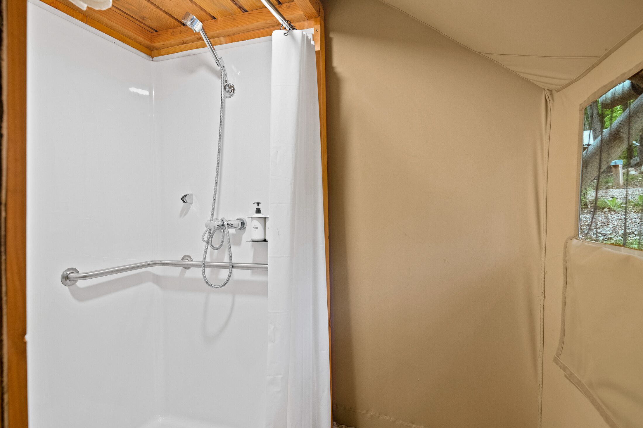 A shower area with a white door, a wooden ceiling, and a window with a view of greenery.