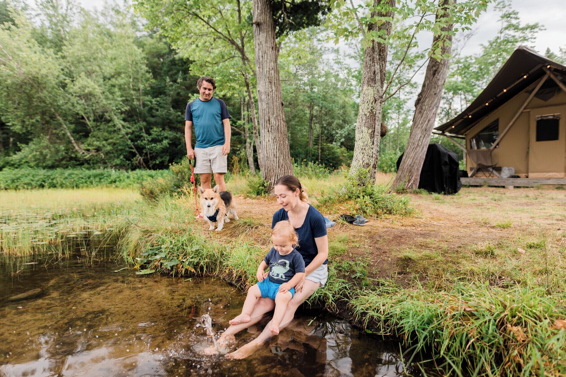 En familie ved en bæk i et skovområde med en hytte i baggrunden.