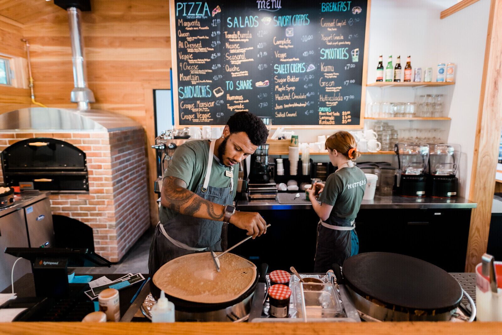 A pizza restaurant with a menu board, a brick oven, and a counter with a person preparing food.