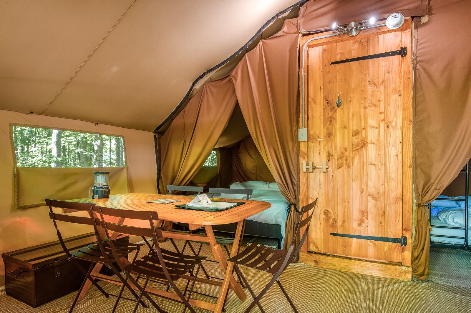 A cozy tented room with a wooden table, chairs, a bed, and a window overlooking a forest.