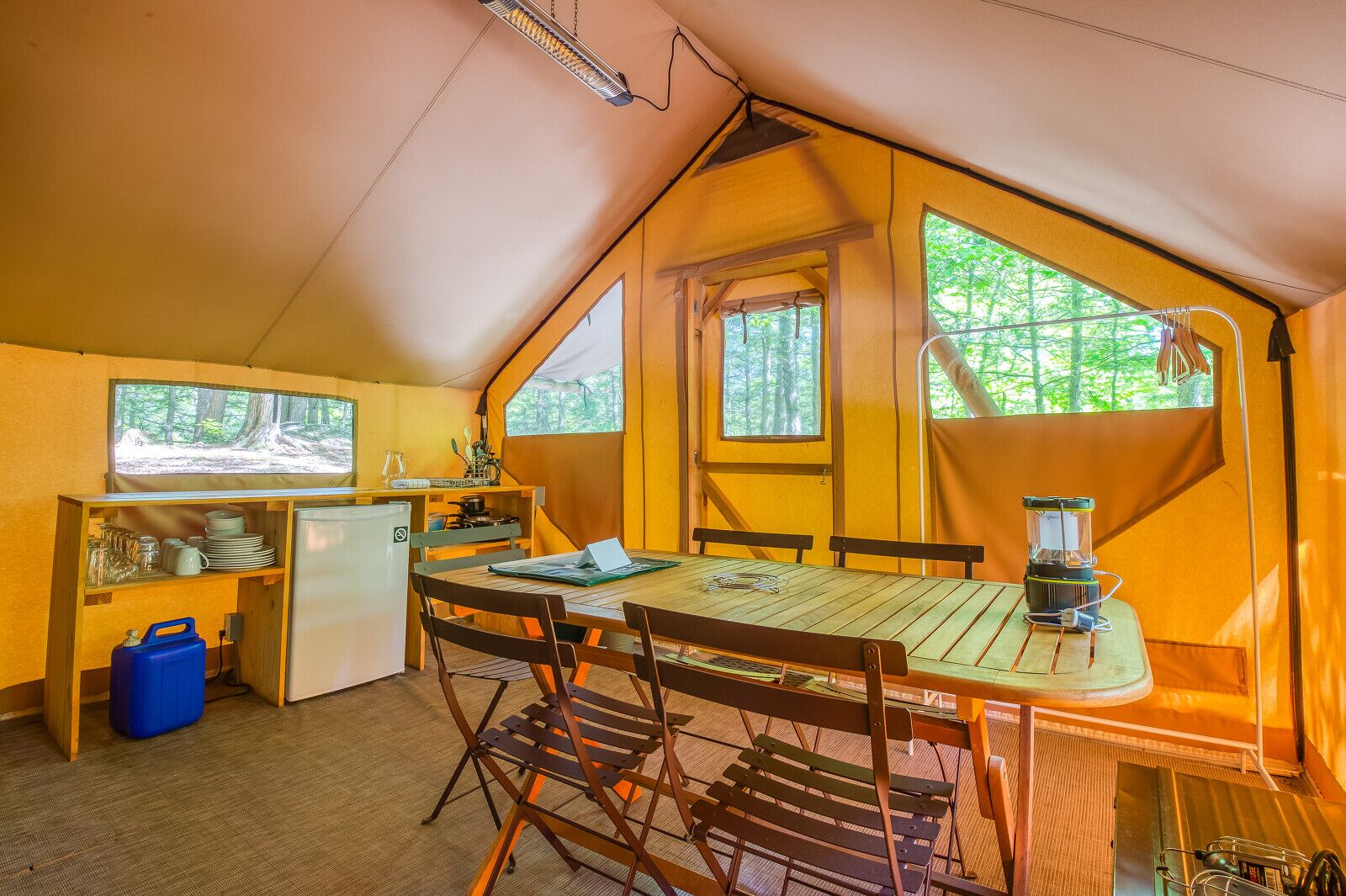 A cozy tented cabin with a kitchenette, dining area, and large windows offering forest views.