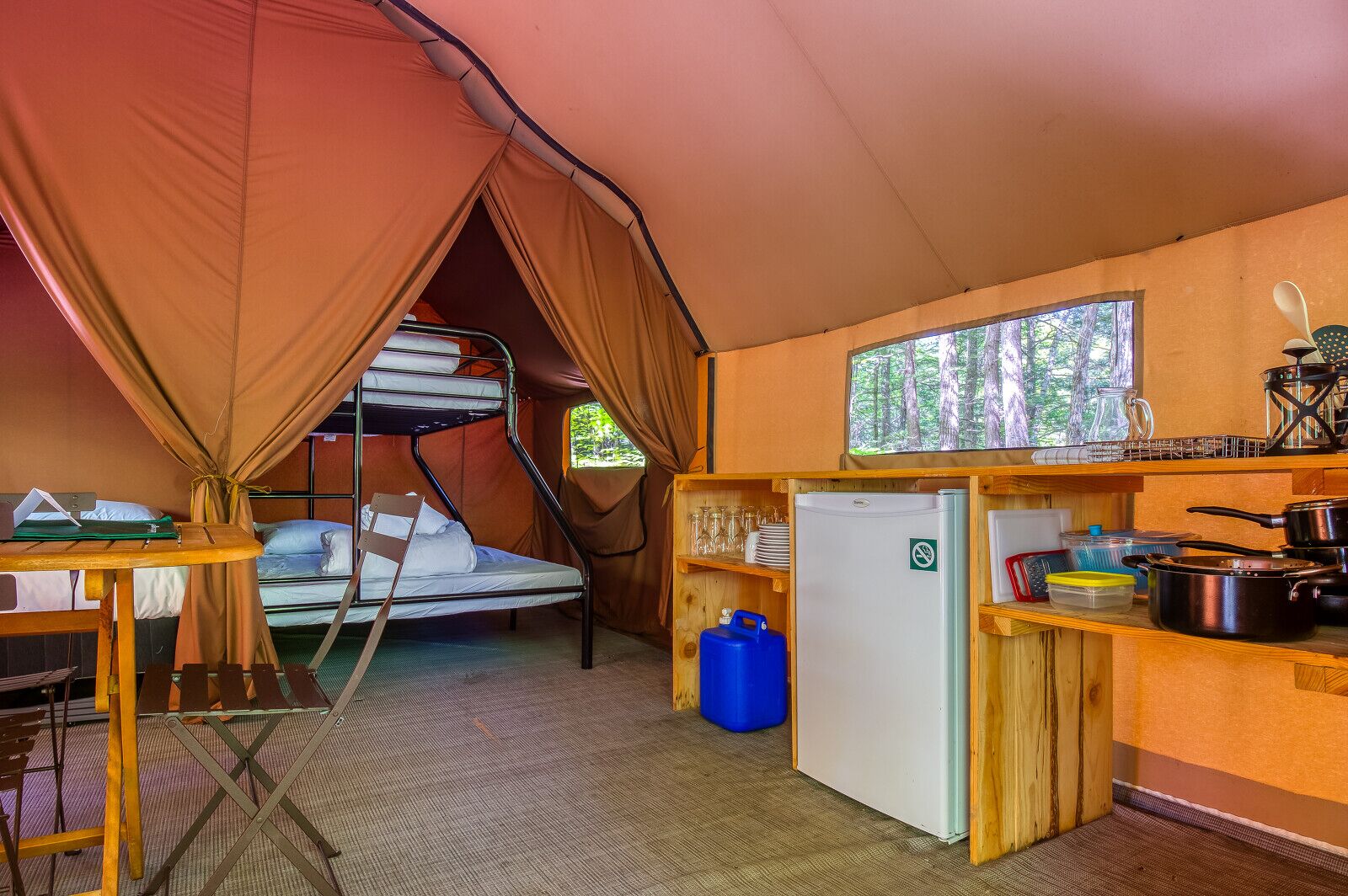Ein kompaktes, gut ausgestattetes Zelt mit Etagenbetten, einer kleinen Küchenzeile und einem Fenster mit Blick auf einen Wald.