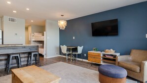 Una sala de estar moderna con una mesa de madera, un reposapiés azul, un sofá color beige, un televisor montado en la pared y una araña.