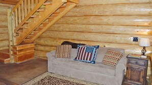 Interior de una cabaña de madera con una escalera de madera, un sofá color beige con almohadones con la bandera estadounidense, una alfombra estampada y un pequeño armario de madera con una lámpara.