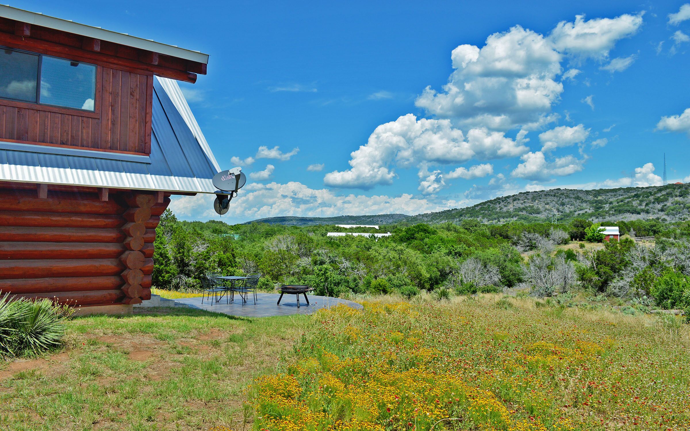 A log cabin with a satellite dish, a patio with outdoor furniture, and a view of a hilly landscape with scattered buildings.