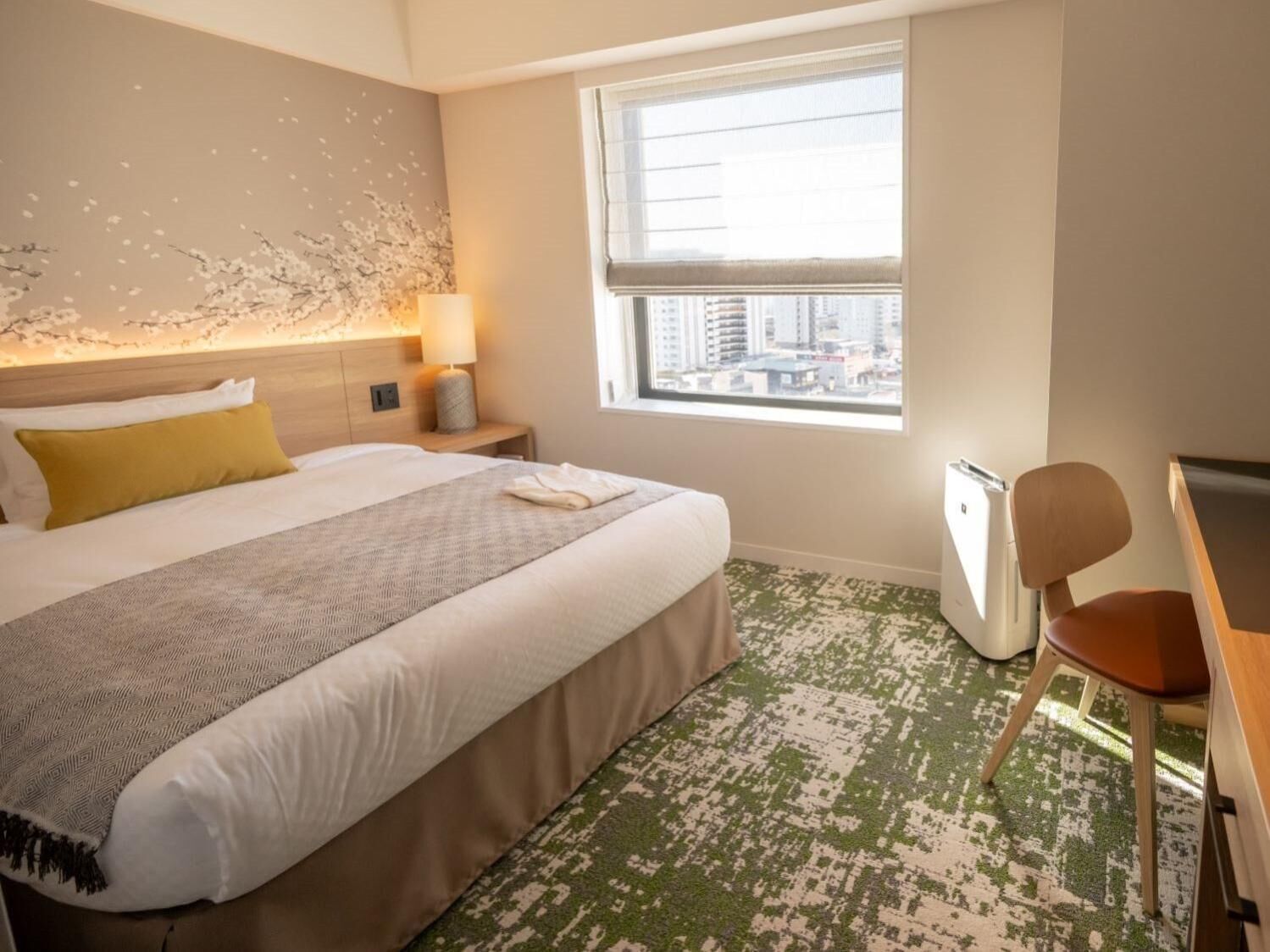 A hotel room with a bed, a desk, a chair, a window with a city view, and a wall with a textured design.