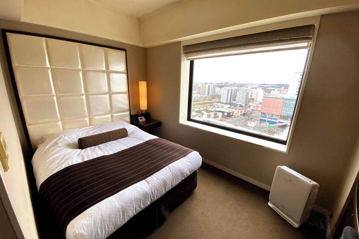 A hotel room with a bed, a nightstand, a lamp, a window with a city view, and a radiator.