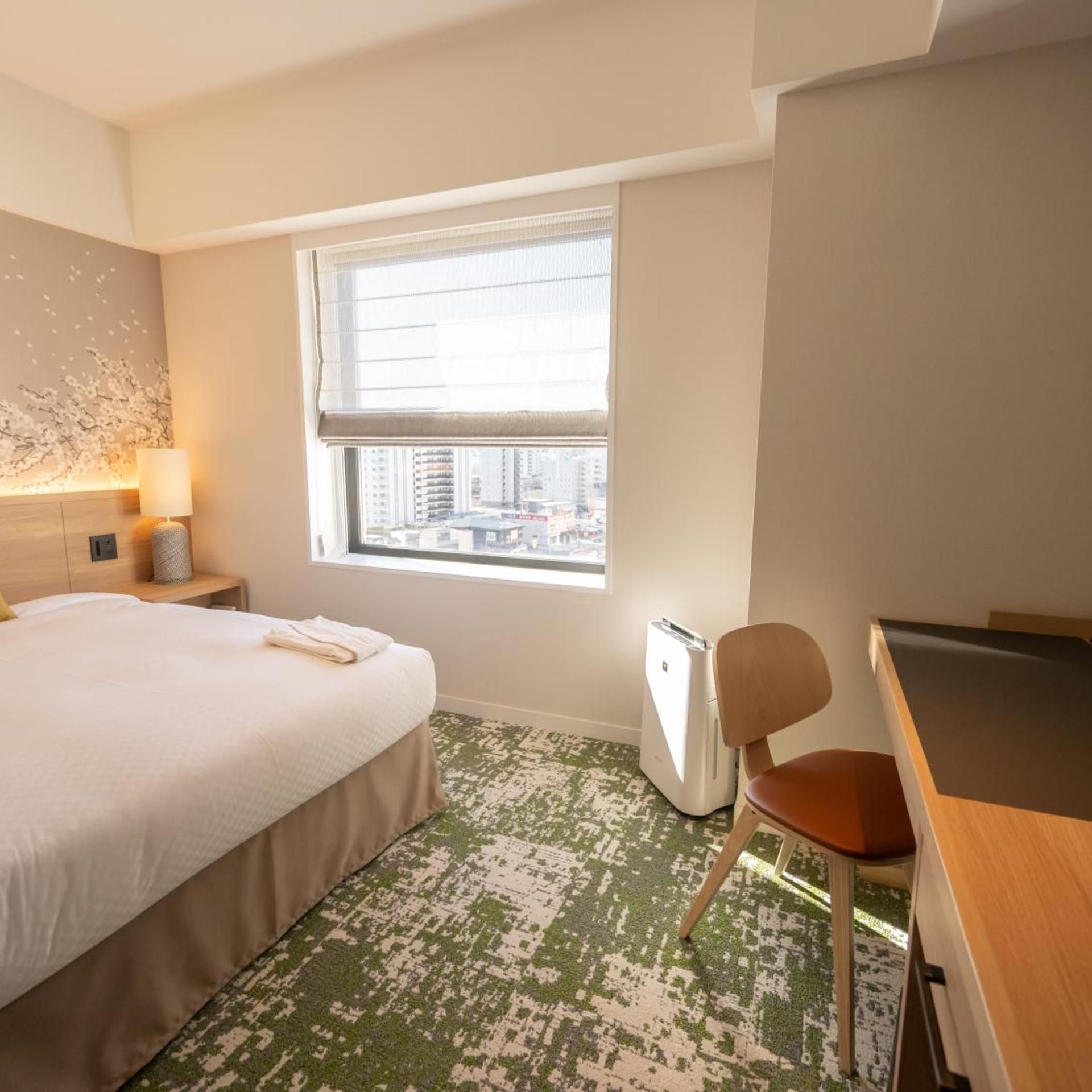 A hotel room with a bed, a desk, a chair, a lamp, and a window with a city view.