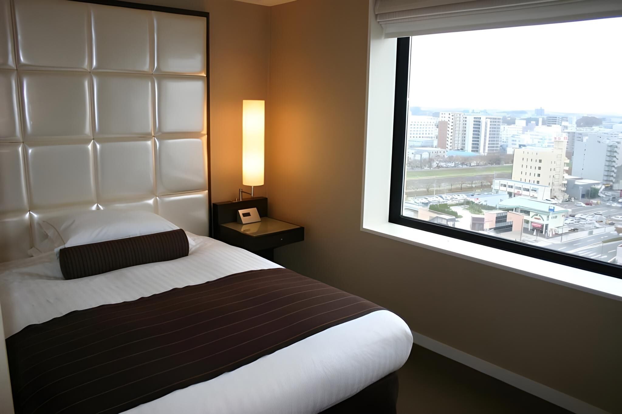 A hotel room with a large bed, a nightstand with a lamp, and a window offering a city view.
