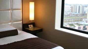 Ett hotellrum med en säng, ett nattduksbord med en lampa och ett fönster med utsikt över staden.