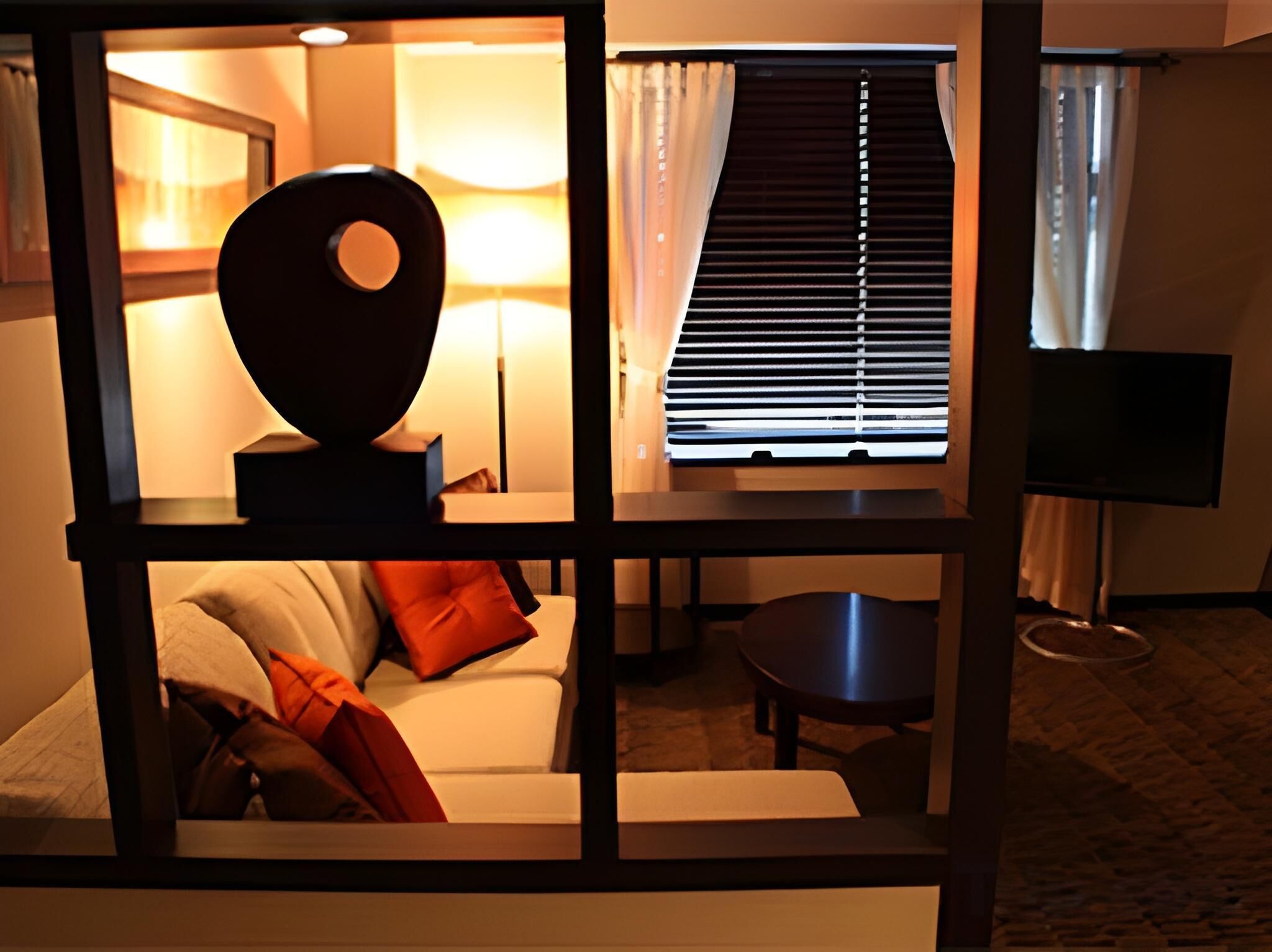 Habitación de hotel con sofá, una mesa, lámpara, televisión y ventana con persianas.