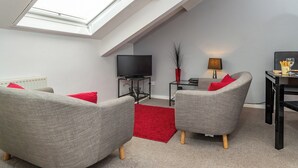 Een moderne woonkamer met dakraam, grijze banken met rode kussens, een flatscreen televisie, een klein bijzettafeltje met een lamp en een rood vloerkleed.