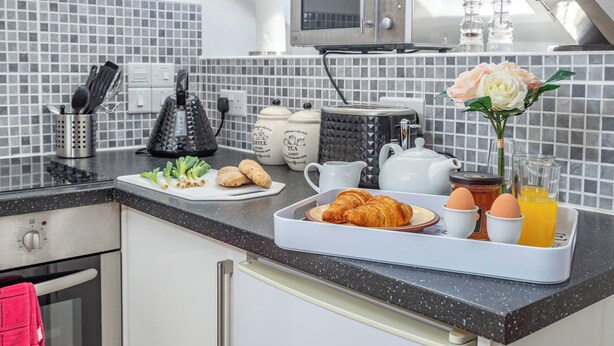 Een moderne keuken met een zwart aanrecht, een magnetron, een waterkoker en een schaal met croissants, eieren en sap.