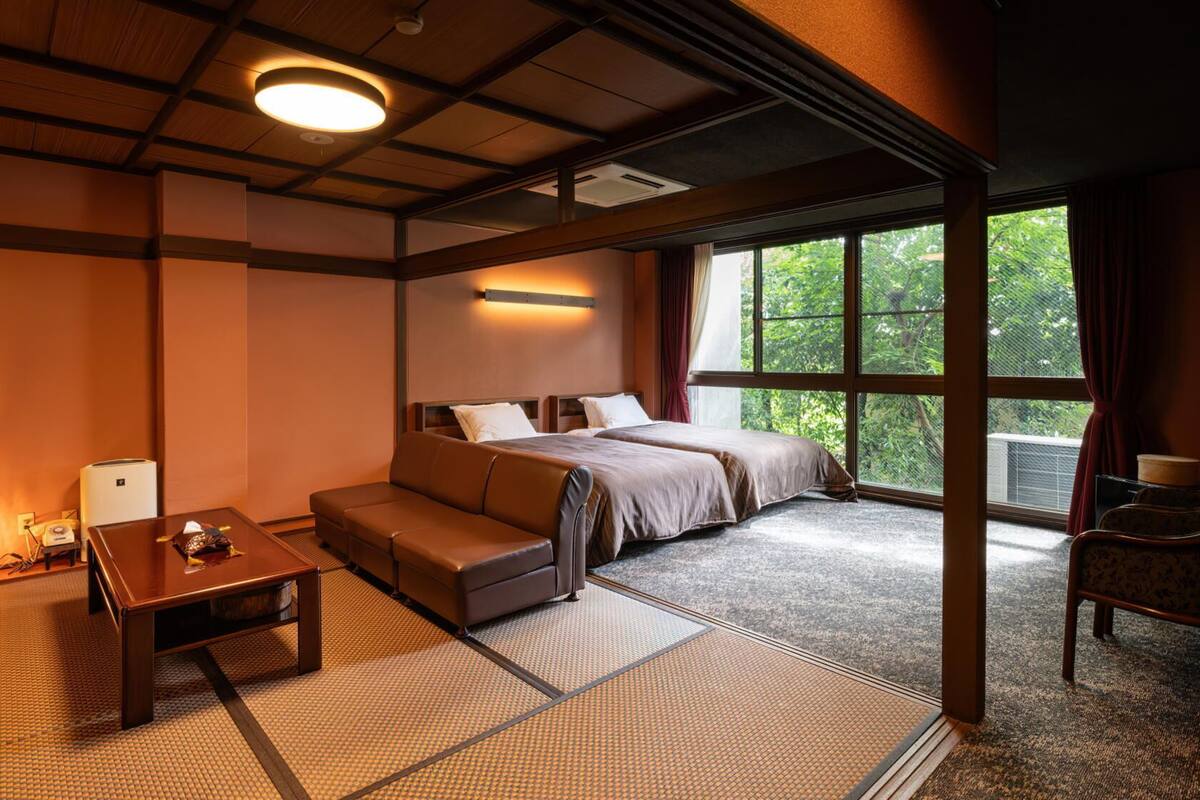 A hotel room with a bed, sofa, and a small table. There is a window with a view of greenery.