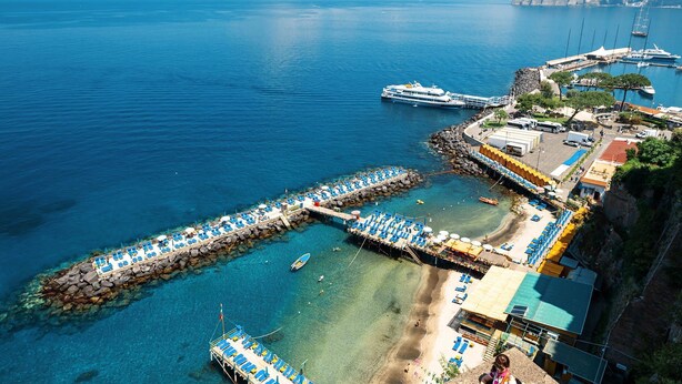 An aerial view of a coastal area with clear blue waters, a pier with blue lounge chairs, a cruise ship docked, and a mountainous backdrop.