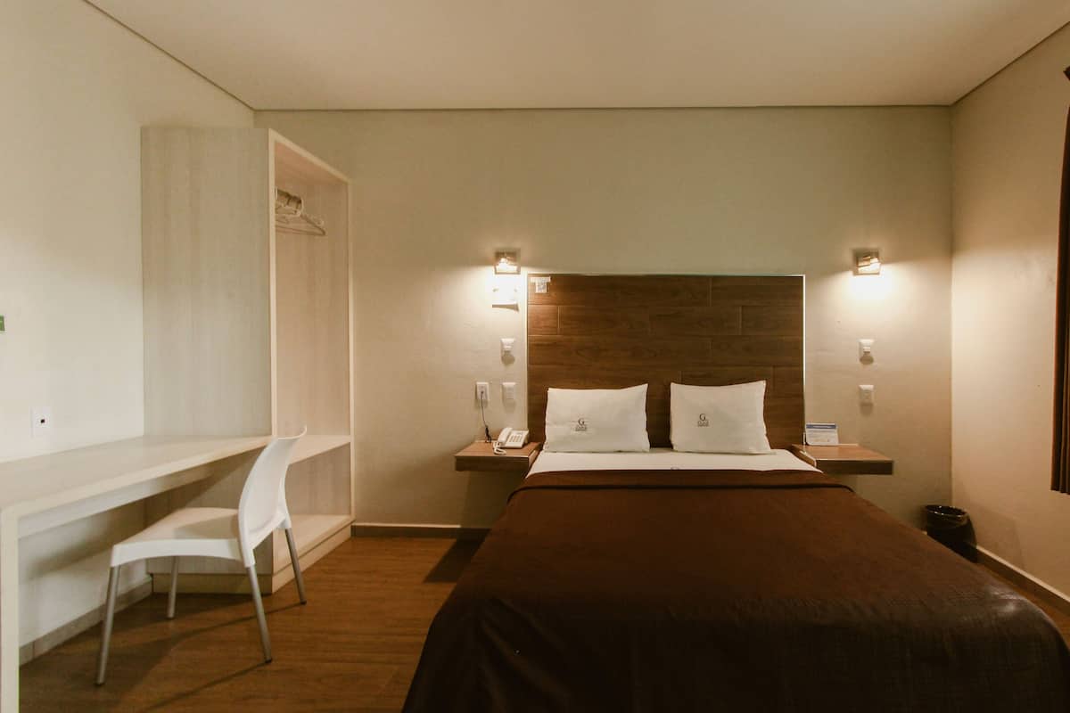 A hotel room with a bed, bedside table, lamp, and a desk with a chair.
