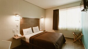 Une chambre d’hôtel avec une tête de lit en bois, un lit recouvert d’une couverture marron, une table de chevet avec un téléphone et une fenêtre avec des rideaux.