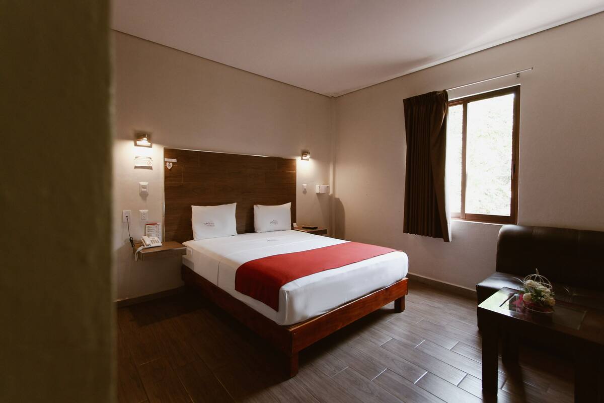 A hotel room with a wooden bed, bedside lamps, a small table with a vase, and a window with curtains.
