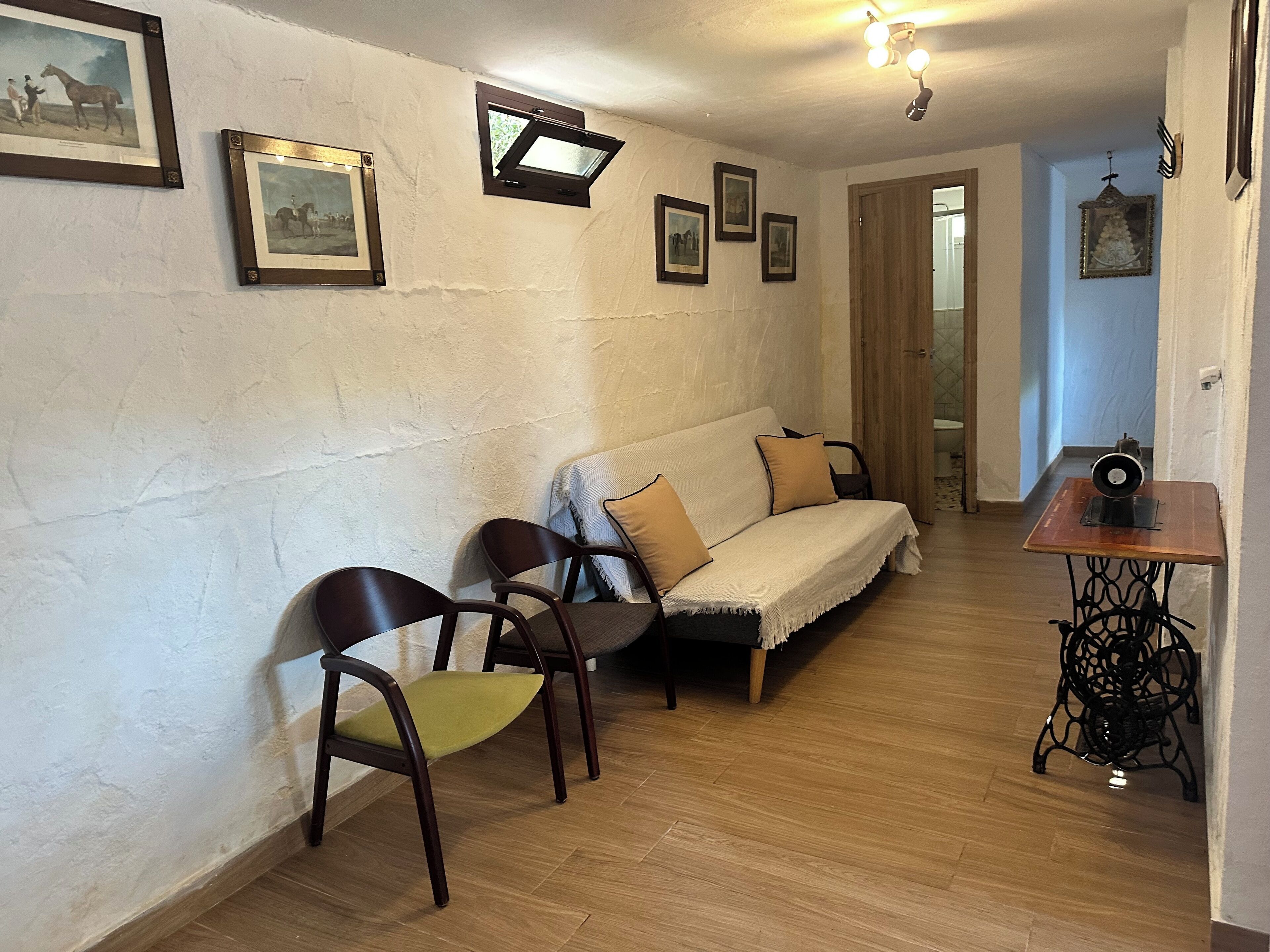 Una habitación con piso de madera, un sofá, dos sillas, una máquina de coser y varias fotografías enmarcadas en la pared.
