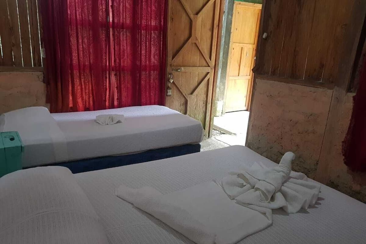 A room with two beds, one with a mattress and pillows, and the other with folded blankets. There is a wooden door and a window with red curtains.